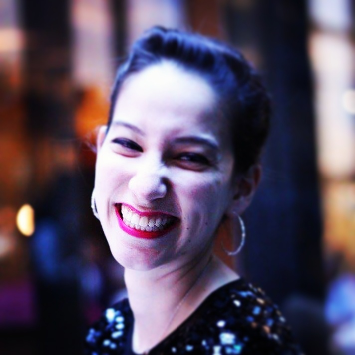 Photo: a young woman with dark hair wearing silver hoop earrings smiles at the camera. She is visible from the neck up. The background is blurry.